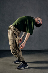 Graceful guy with tattooed body, earrings, beard. Dressed in khaki t-shirt, overalls, black sneakers. Dancing on gray background. Dancehall, hip-hop