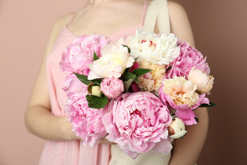 Woman with bouquet of beautiful peonies in bag on beige background, closeup
