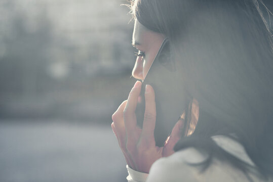 Mobile Conversation. Side View Close Up Of A Young Beautiful Woman Which Engaged In A Phone Call, Holding A Modern Smartphone To Her Ear In A Naturally Lit Outdoor Environment.