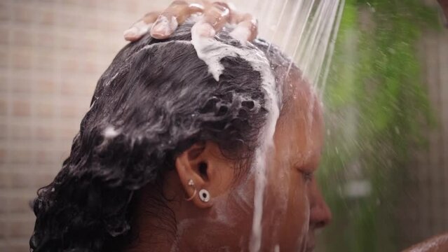 Close-up Of Black Woman Face Washing And Rinsing Her Curly Hair With Water In Shower. African American Young Female Using Shampoo For Hygiene. Daily Personal Body Care In Bathroom At Home. Slow Motion