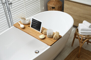 Wooden tray with tablet and spa products on bath tub in bathroom