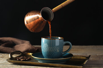 Turkish coffee. Pouring brewed beverage from cezve into cup at wooden table against black background