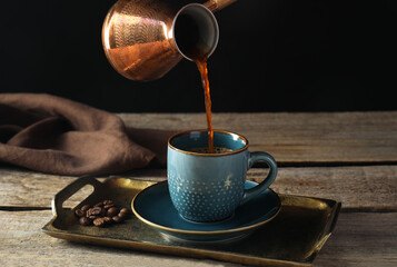 Turkish coffee. Pouring brewed beverage from cezve into cup at wooden table against black background
