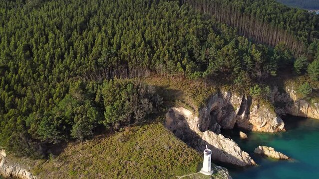 Picturesque landscape of northern Spain. Lighthouse isolated on the rocks, surrounded by forest and hills. In front of them the sea in turquoise colors. Drone backwards ascending.