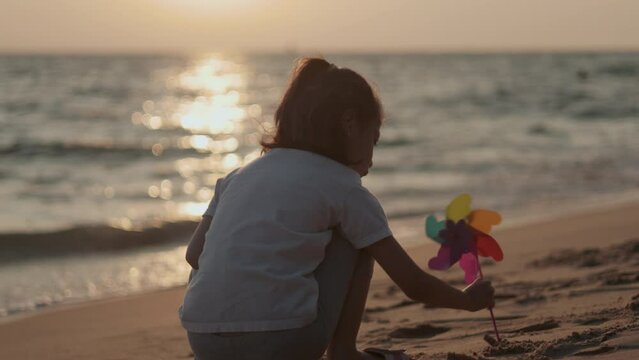 Adorable Asian kid who is playing sand on the beach under beautiful sky, coastline, horizon and sea at the sunset time with carefree, happiness and enjoyment of children on holiday in summertime.