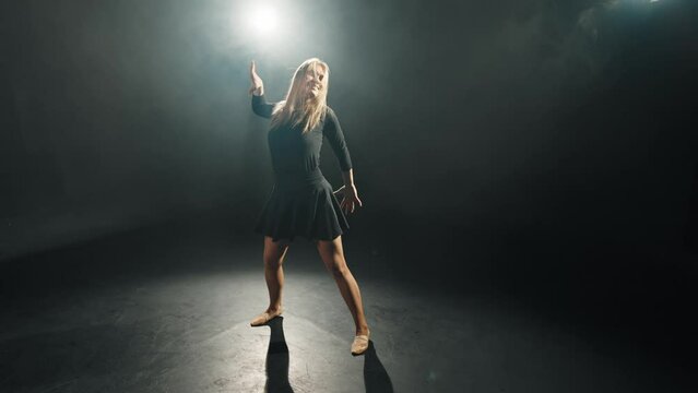 Woman choreographer dancing jumping. Blonde hard-working female ballerina dances in black dress in studio on stage. Performing dance element classical ballet bouncing in air. Classical choreography.
