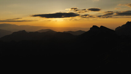 Yellow twilight in the mountains during sunset. The sun is hidden in the clouds. Long black rows of silhouettes of mountain peaks. Epic sunset in the mountains. The beauty of nature.