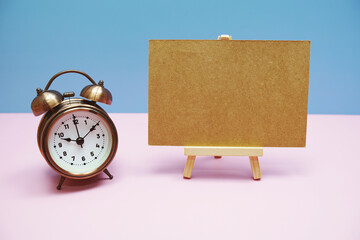 Empty card mockup space for text message with wooden easel and vintage alarm clock on pink...