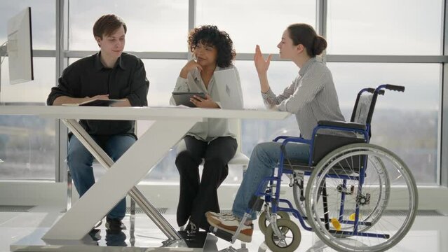 Paralyzed woman works with colleagues. Female in wheelchair has spine legs trauma discussing working project in office on meeting with multiethnic team. Teamwork, brainstorming, special needs person.
