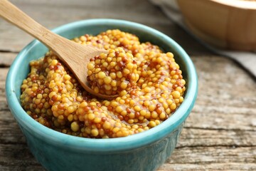 Whole grain mustard in bowl and spoon on wooden table, closeup