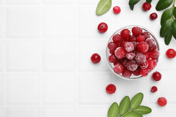 Frozen red cranberries in bowl and green leaves on white table, flat lay. Space for text