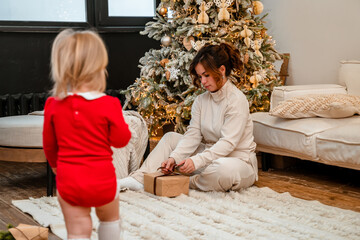 Beautiful brunette woman mother with baby toddler child ,spends the New Year holiday, sitting on a white floor with Christmas tree. Festive family atmosphere.