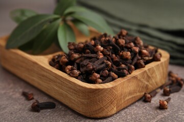 Wooden tray with aromatic cloves and green leaves on brown table, closeup