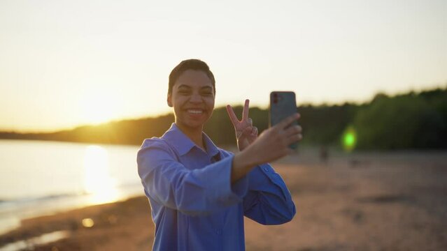Teenager girl makes selfie on sea beach. Indian teen girl with short hair smiling looking at smartphone camera at sunset resting on ocean beach. Taking picture. photographing, social media addiction.