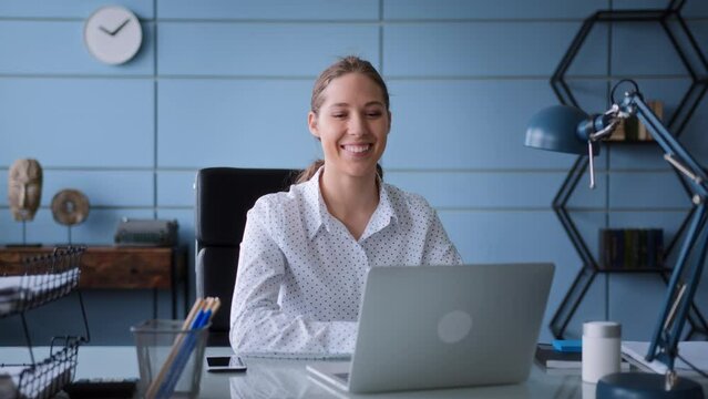 young woman in white shirt sits at table in office with laptop, typing on keyboard concentrated. smiles and rejoices to success result and great news, looking to monitor