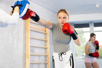 Determined adolescent girl in boxing gloves training with group of teenagers in gym, punching pads...