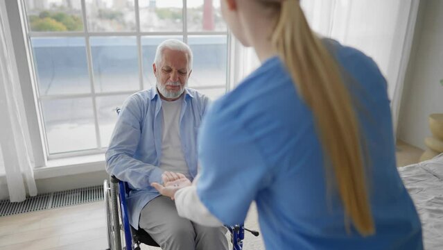 Nurse gives old wheelchair man pills. Physical therapist woman provides medical help support to senior paralyzed male gives water medications. Palliative care of neurodegenerative pathologies concept.