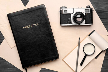 Holy Bible with photo camera, magnifier, notebook and paper sheets on dark wooden background