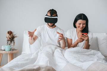 Couple in bed with man in VR headset gesturing and woman using smartphone, amused