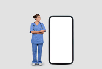 Smiling european nurse in blue uniform and stethoscope, standing beside a giant smartphone