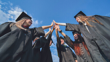 Group of happy successful graduates in academic hats and robes standing in circle and putting their...