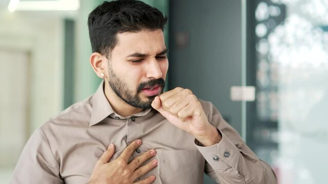 Businessman in a shirt coughing while sitting at workplace at in business office. Sick with a cold or flu employee feels bad, holds his hand to his chest. Bearded male worker has bronchitis. Close up