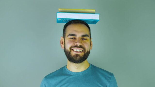 young bearded man with a pile of books on his head pointing up while looking at camera