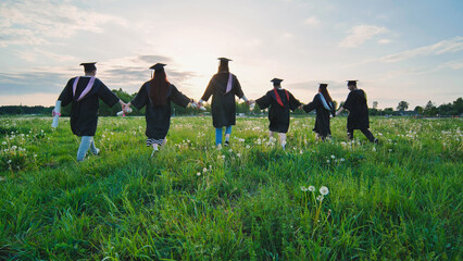 Six graduates in robes walk against the backdrop of the sunset.