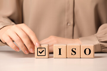 Woman adding cube with check mark to abbreviation ISO of wooden cubes at white table, closeup