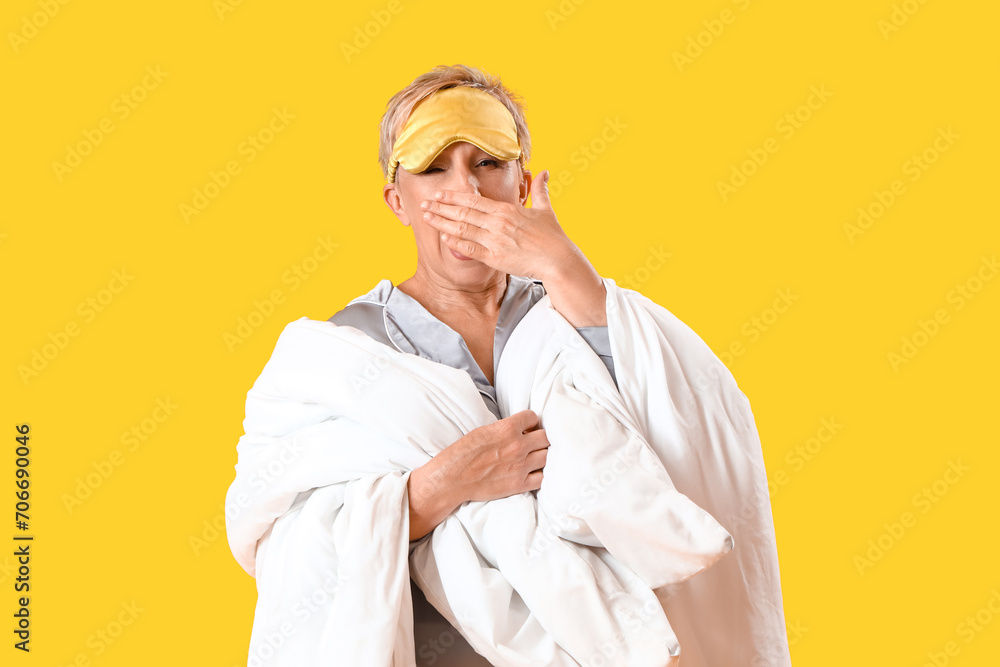 Wall mural Mature woman with sleeping mask and blanket yawning on yellow background