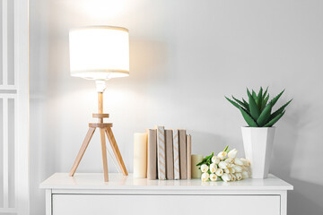 Glowing lamp with books, tulips and houseplant on chest of drawers in room