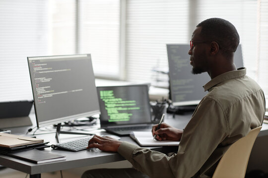 Side view of African American IT specialist typing on computer keyboard and writing in notebook while sitting at desk in cybersecurity office
