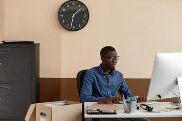 Medium shot of Black man wearing glasses working on computer while sitting at desk in spacious...