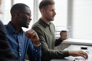 Side view with focus on smiling African American male IT professional wearing glasses working on code with Caucasian colleague while sitting at table in office