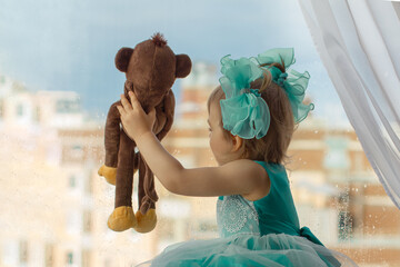 little girl of three years with blue bows and dress looks with monkey toy out window