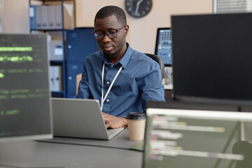 Medium shot of focused African American male programmer in glasses typing on laptop while sitting at desk, look through computer monitors in blurred foreground