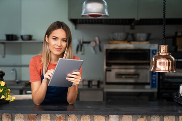 Beautiful confident young woman wearing apron looking at camera holding digital tablet fintech...