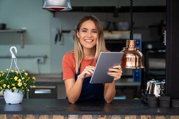 Smiling confident young woman wearing apron looking at camera holding digital tablet fintech device...