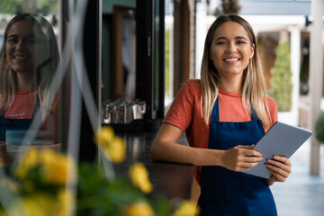 Smiling confident young woman wearing apron looking at camera holding digital tablet fintech device standing outside the restaurant or Cafe. - obrazy, fototapety, plakaty