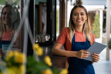 Gordijnen Smiling confident young woman wearing apron looking at camera holding digital tablet fintech device standing outside the restaurant or Cafe. © Dorde