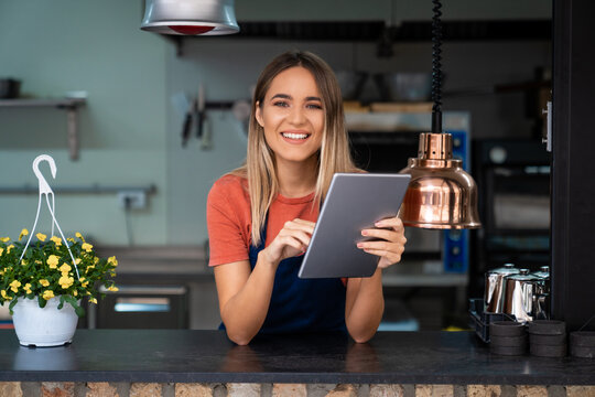 Smiling confident young woman wearing apron looking at camera holding digital tablet fintech device standing in commercial kitchen. Female small bakery owner managing her business using digital tablet