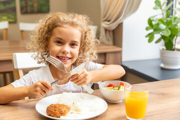 Little cute child sitting at table with plate of food and glass of orange juice. Kid have a meal at home. Caucasian toddler girl have meat, fresh vegetables with rice lunch, healthy nutrition concept