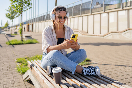 hipster woman in bluetooth headphones enjoying positive music playlist from mobile radio using 4g wireless in city, have video call, girl holding coffee to go and smartphone.