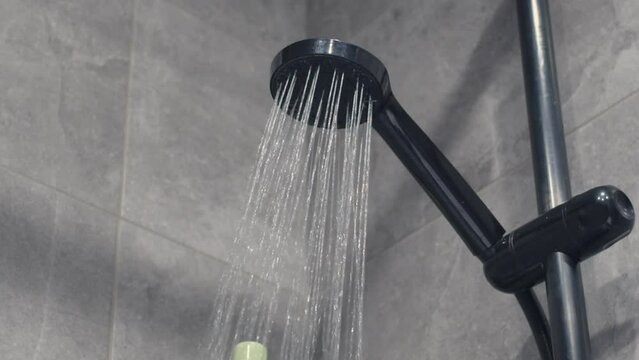 Water pours from a black shower head in slow motion against the background of gray tiles and shampoo bottles standing on the shelf.
