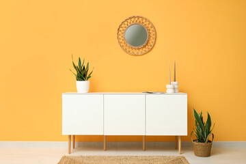 Modern white chest of drawers with houseplants and mirror near orange wall in room