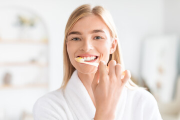 Portrait of pretty blonde woman attentively cleaning her teeth indoors - obrazy, fototapety, plakaty