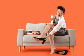 Young pin-up businesswoman with coffee and folders taking off heels on sofa against orange background - obrazy, fototapety, plakaty