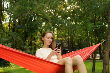 A young smiling woman sits on an orange hammock attached to a tree on a green background in spring or summer and looks at the phone. The concept of outdoor recreation, social networks