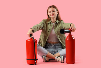 Young woman with fire extinguishers sitting against pink background