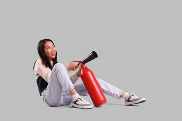 Young Asian woman with fire extinguisher sitting against grey background